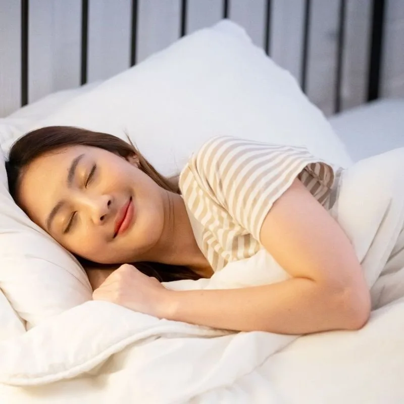 A woman resting on her side in bed in soft morning light, eyes closed, calm under a white duvet