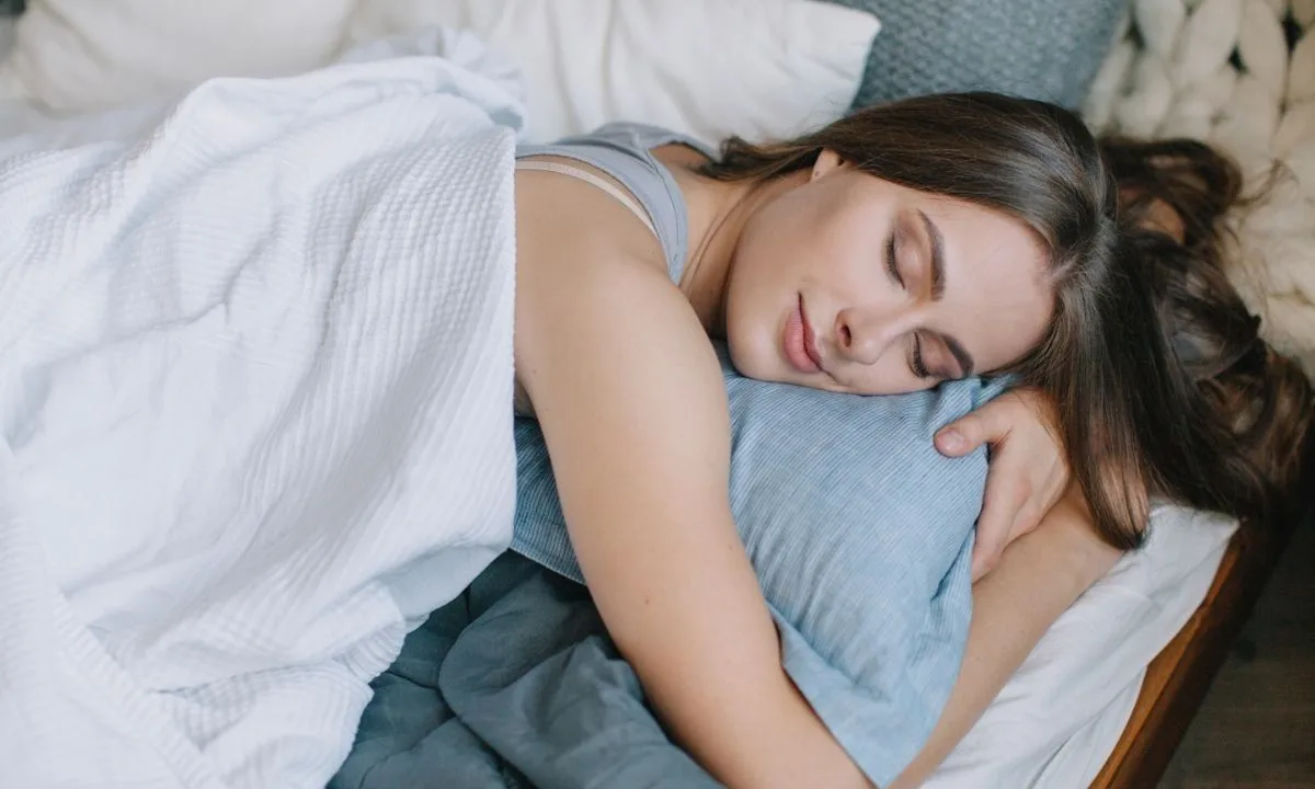 A woman sleeping peacefully on her side in a softly lit bedroom, one hand resting on the pillow beside her face